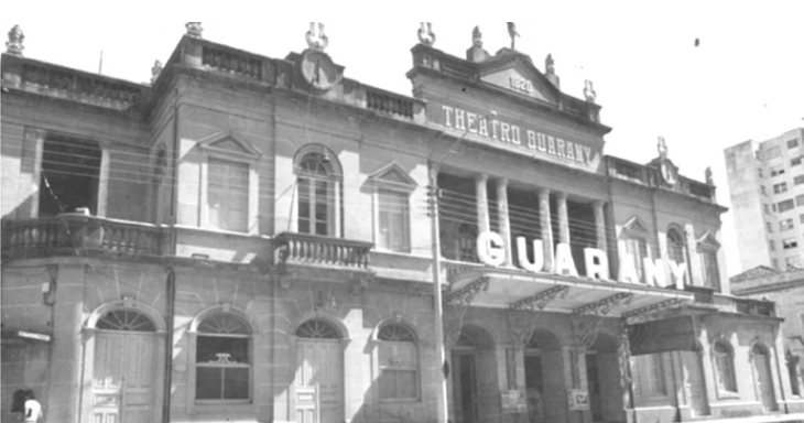Fotografia histórica em preto e branco do Theatro Guarany com letreiro visível no frontão, arquitetura eclética com colunas, balaustradas e frontões triangulares, edifícios modernos ao fundo indicando transformação urbana, documentando este importante cinema que integrava o circuito de entretenimento do centro histórico de Pelotas