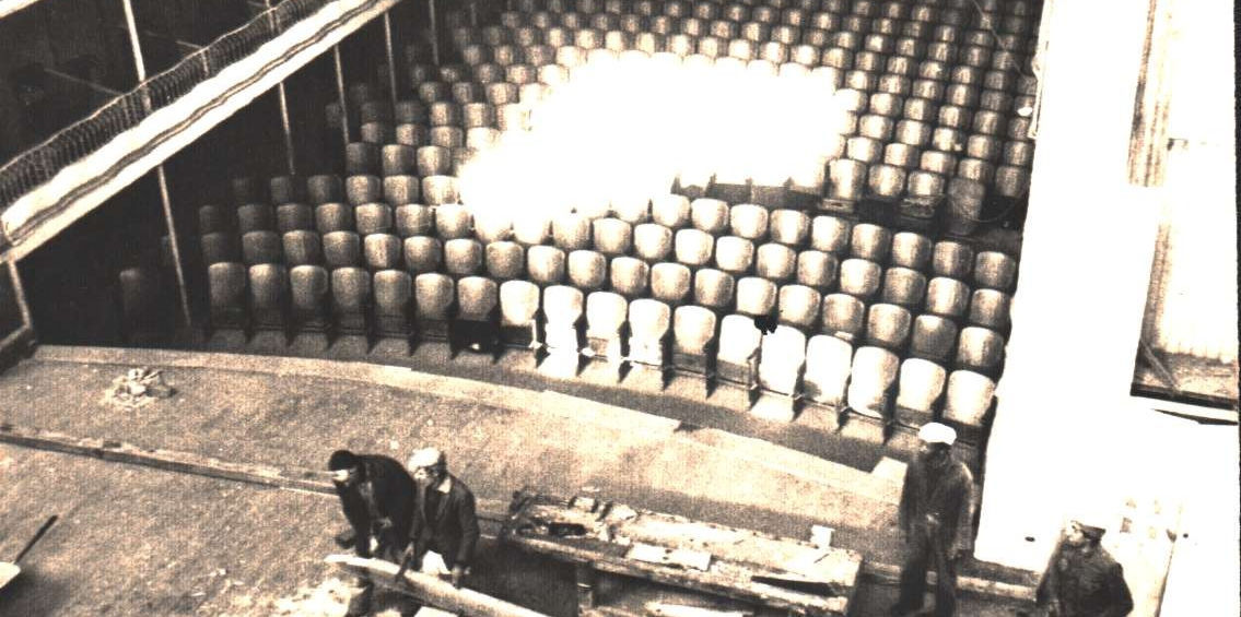 Fotografia histórica em preto e branco do interior do Theatro Sete de Abril mostrando plateia em formato semicircular com fileiras de poltronas dispostas em degraus, pessoas trabalhando na montagem de elementos cenográficos no palco, documentando tanto a arquitetura interna quanto os bastidores da produção teatral neste importante espaço cultural pelotense mapeado pelo projeto Inventário de Plateias