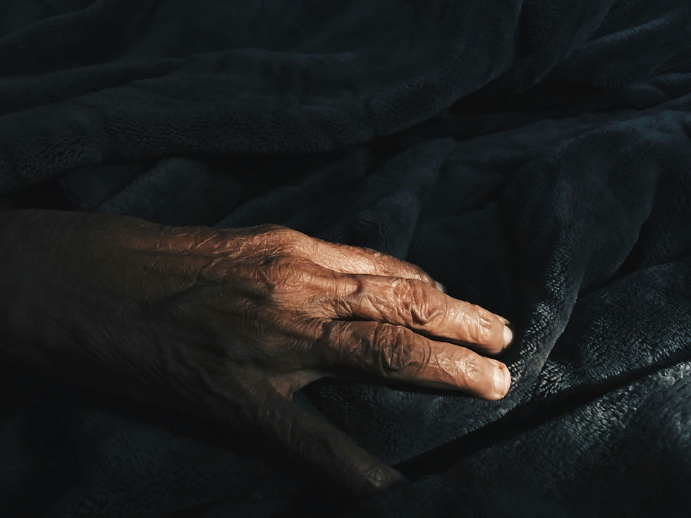 A wrinkled hand rests gently on a soft, dark blanket in low light, evoking a sense of calm and introspection.