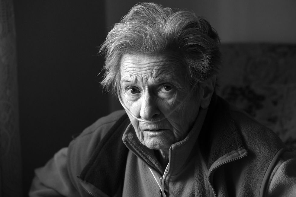 Elderly person with oxygen tube, wearing a jacket, looks pensively at camera. Black and white setting with a soft, dim background.