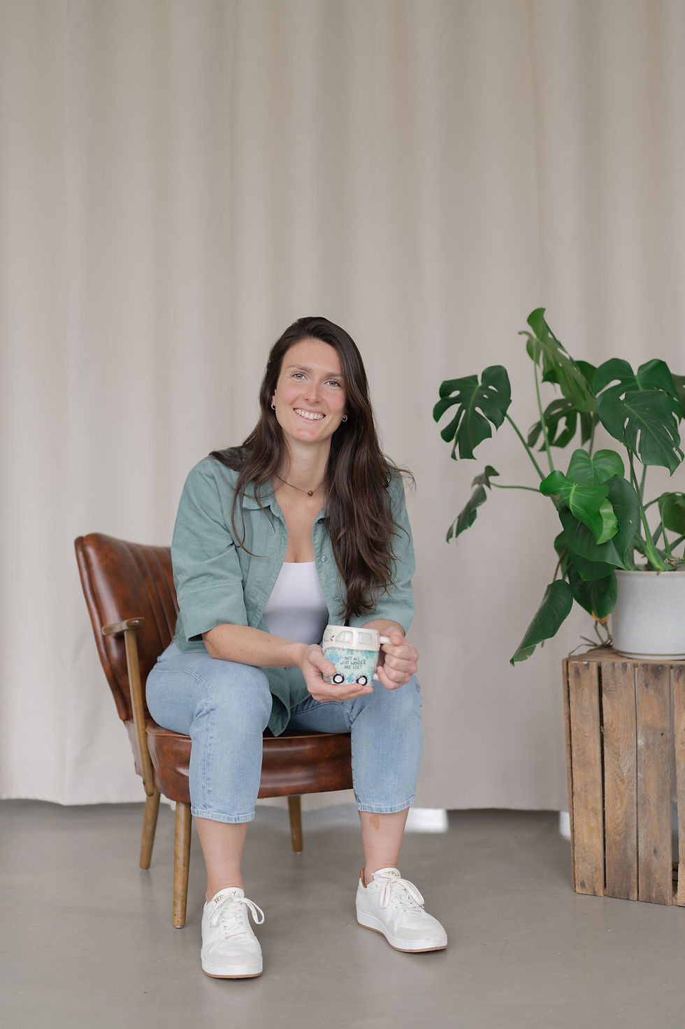 Virtuelle Assistenz Vivien Schröder sitzt auf einem braunen Ledersessel, daneben steht eine alte Weinkiste mit einer Monstera Grünpflanze drauf, im Hintergrund ist ein warm-beiger Vorhang. Die selbstständige Frau hat sich von Christiane Timm Brand Fotografie & Markenstrategie, einer Personal Brand Fotografin in Lüneburg und Hamburg, im Rahmen einer Mini Brand Shooting Experience markenstrategisch fotografieren lassen im Studio Tulipan Hamburg.