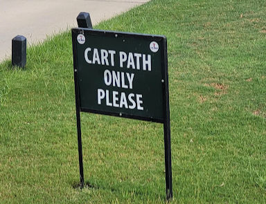 Sign reading "CART PATH ONLY PLEASE" on a grassy area by a paved path, with a calm outdoor setting.