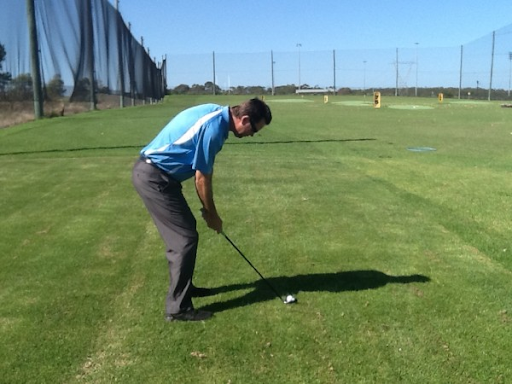 Man in blue shirt prepares to hit a golf ball on a green field. Clear blue sky, netted fence on the side. Calm and focused mood.
