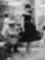 Woman in a black dress poses while Coco Chanel adjusts the hem in a fitting room. Elegant setting, mirrors in the background. Vintage vibe.