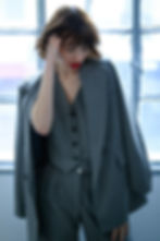 Woman in a grey pinstripe power suit adjusts her hair, with red lips. She stands in front of a large window, creating a moody atmosphere.