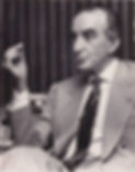 Black-and-white portrait of an older man in a suit and patterned tie, seated and gesturing with raised hand before dark curtains.
