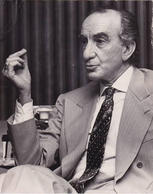 Black-and-white portrait of an older man in a suit and patterned tie, seated and gesturing with raised hand before dark curtains.