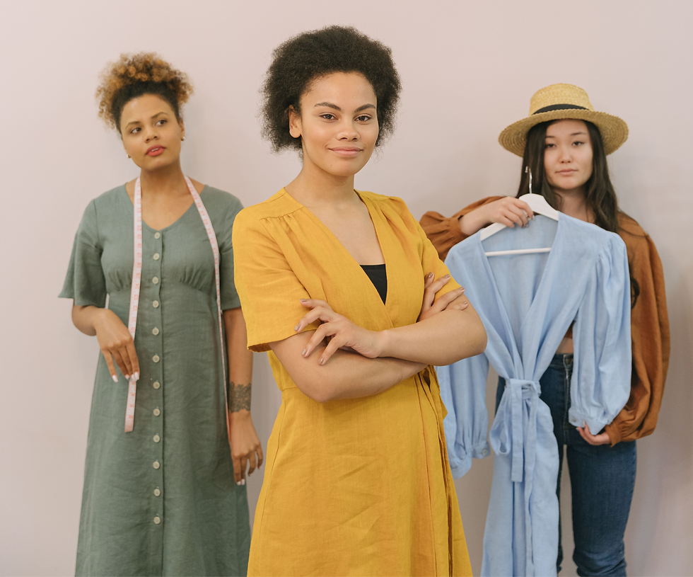 Three women pose in a studio: one in a yellow dress smiles centre, another with a tape measure, and one in a straw hat holds a blue blouse.