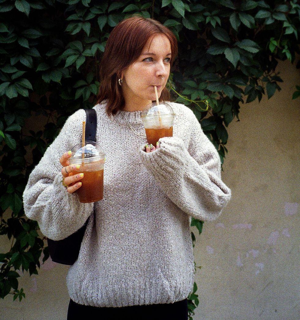 Woman wearing an oversized grey knitted jumper as part of a relaxed everyday outfit. Carrying 2 iced teas and drinking through one of the straws.