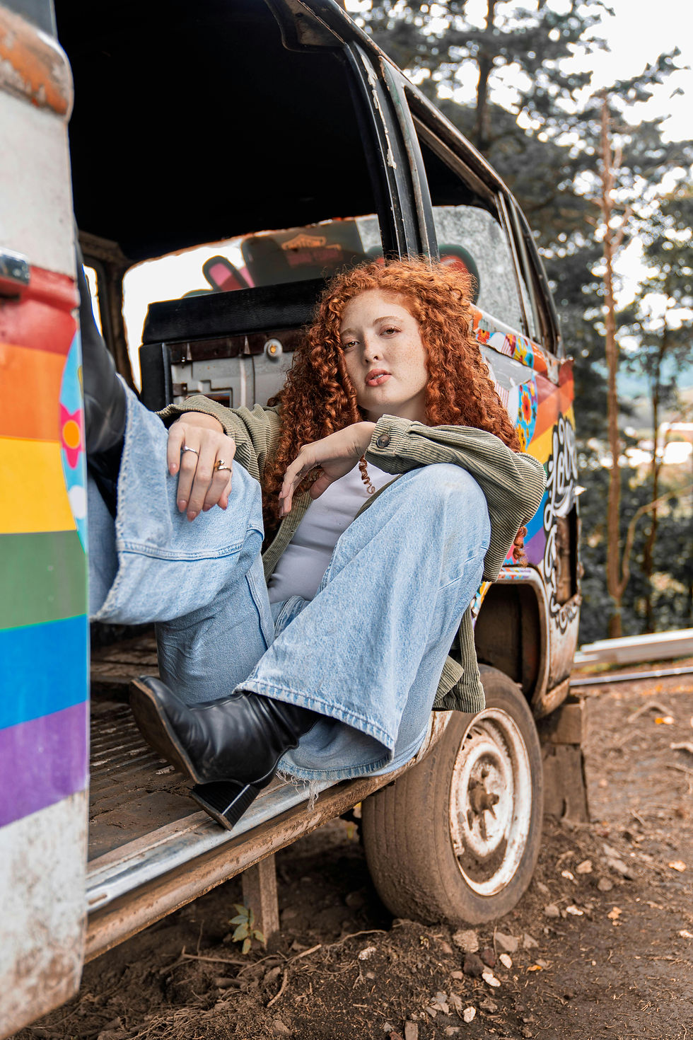 Red-haired person in a jacket and jeans sits casually at the door of a colorful van with rainbow stripes, set in a wooded area. Moody vibe.
