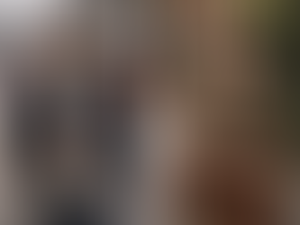 Man with coffee cup and woman with handbag, both wearing tweed caps and jackets. Set in an urban street with double-decker buses.