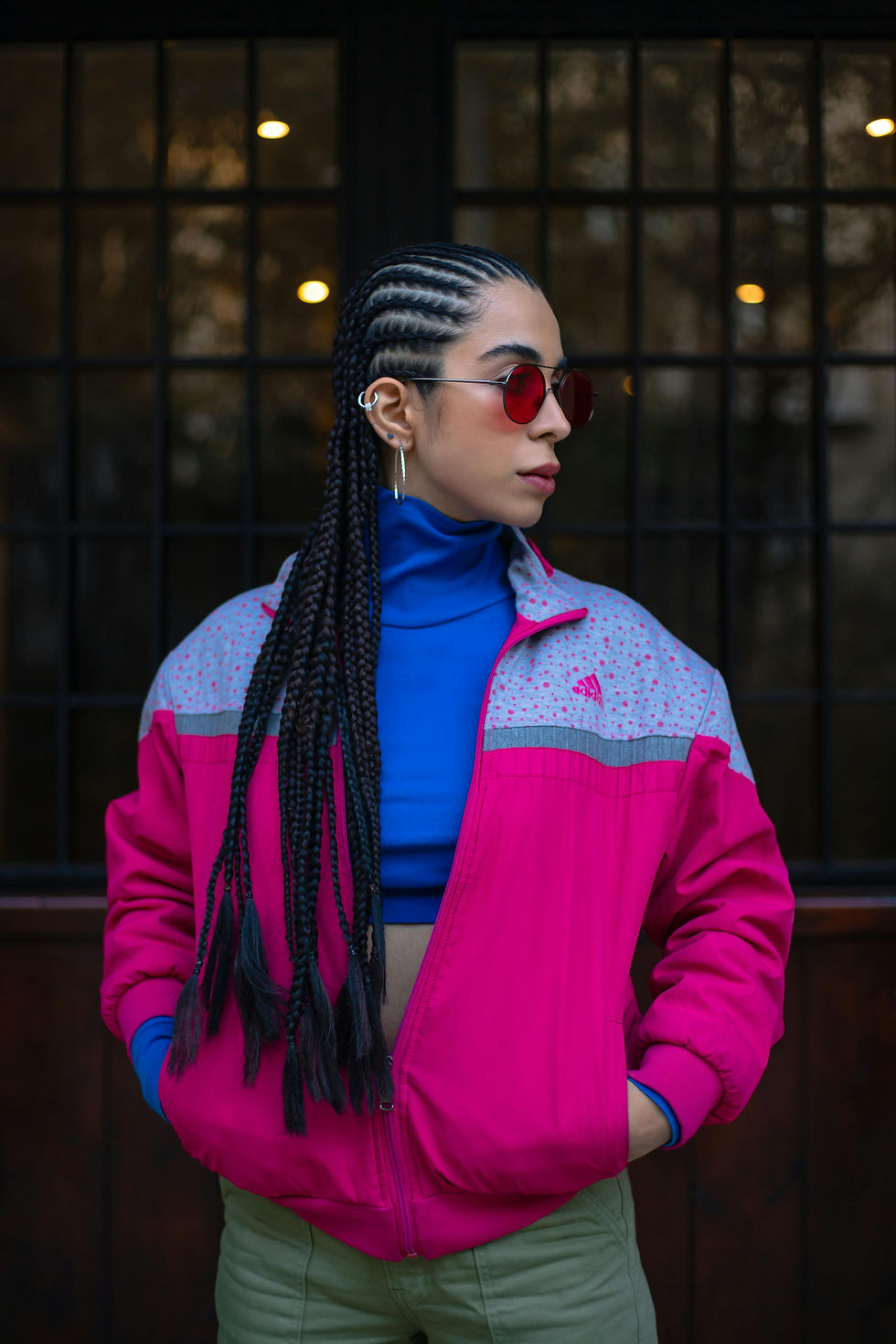 A person with braided hair wears red sunglasses, a pink and lilac retro track jacket by Adidas and a blue top> They are standing against a dark windowed background.