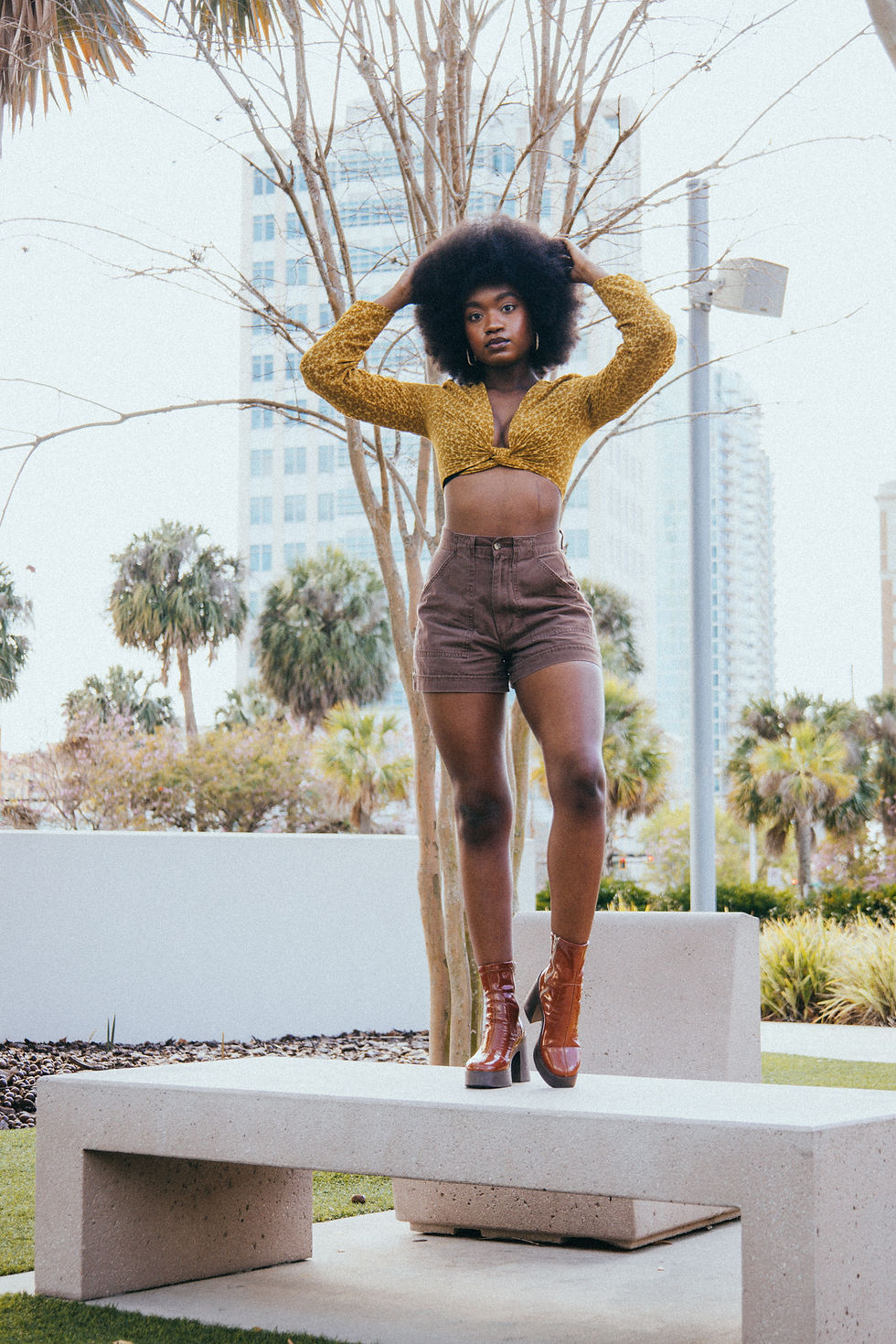 Woman posing on a concrete bench in a city park, wearing a gold crop top, brown shorts and vintage platform boots, with trees and high-rises behind her.