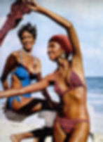 Two women in bikinis smiling at the beach; one in a blue suit, the other in striped, with a matching headscarf. Sunny and cheerful mood. 1970s photo.