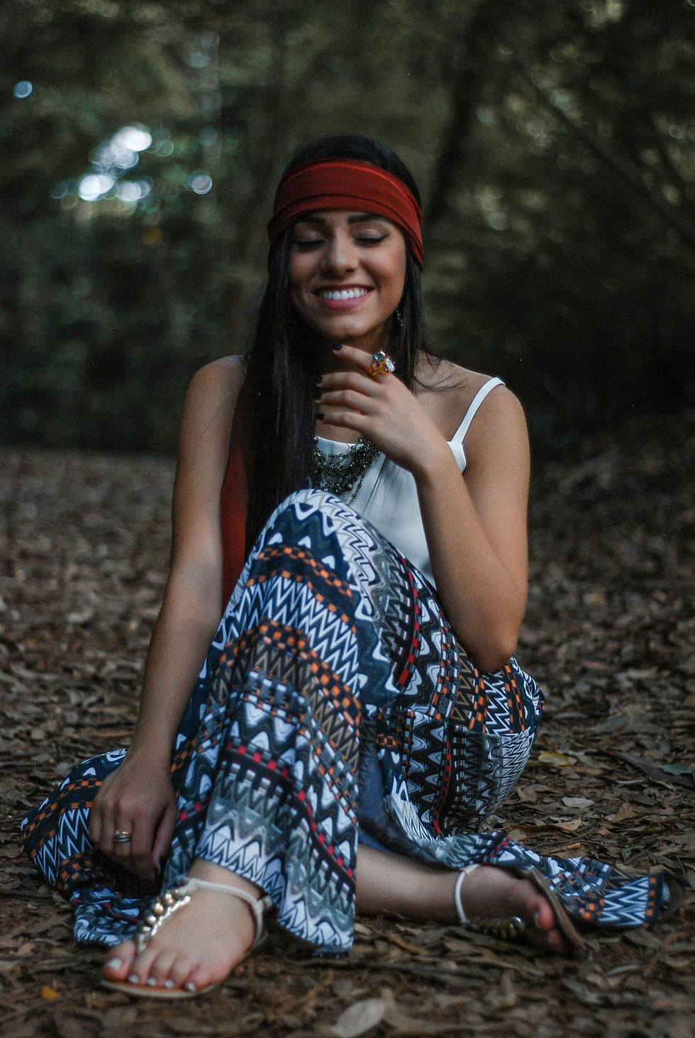 Smiling woman with long hair and red headband sits in forest wearing patterned skirt. Leaf-covered ground, relaxed mood, no visible text.
