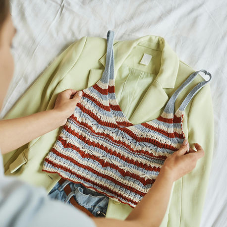 Person holds a striped vintage crochet top over a light yellow blazer on a bed. Neutral tones, casual atmosphere, no visible text.