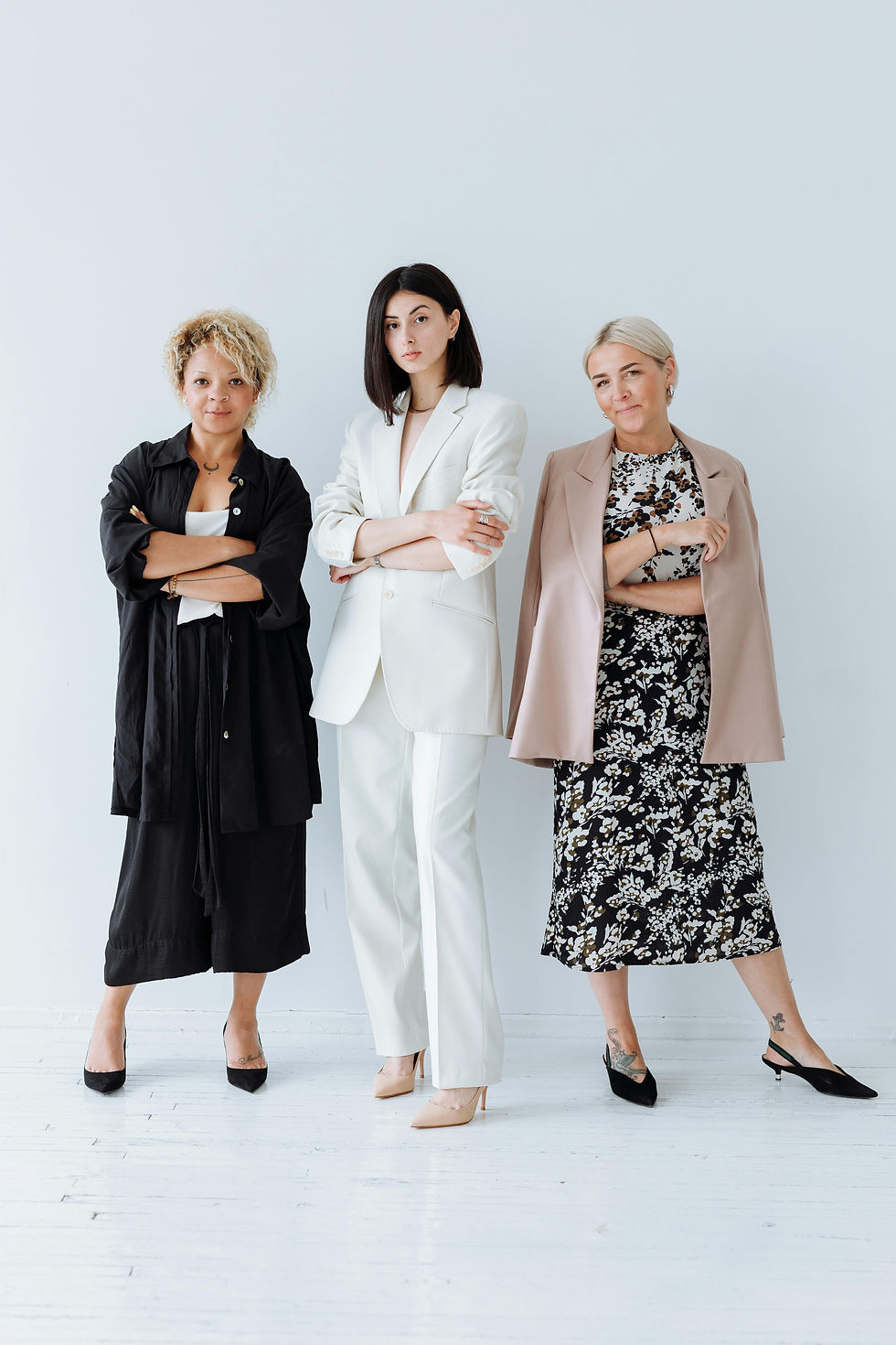Three women stand with crossed arms in a bright studio, wearing black, white, and beige outfits, looking confident.