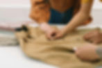 Hands inspect a tan button-up vintage shirt on a table beside neatly folded clothes in a bright sewing or retail workspace.