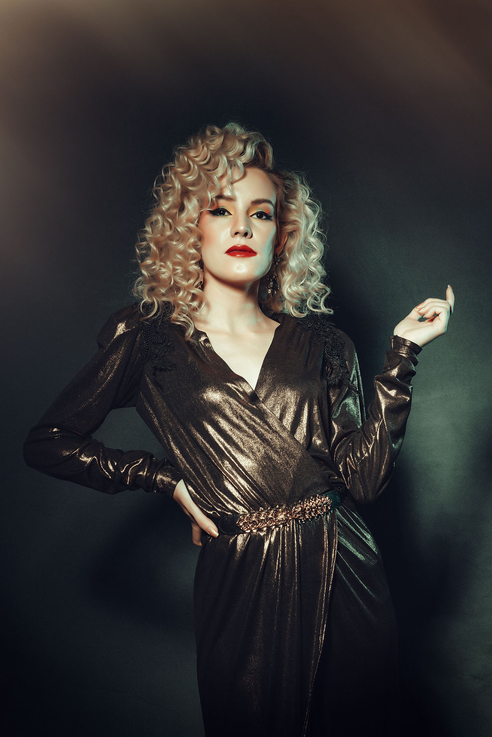 Glamorous blonde woman in a metallic bronze dress poses against a dark studio backdrop, red lipstick and curled hair, confident.