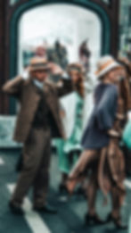 People in 1920s vintage style attire, including hats, dance joyfully on a city street near a shop window. Background features mannequins.