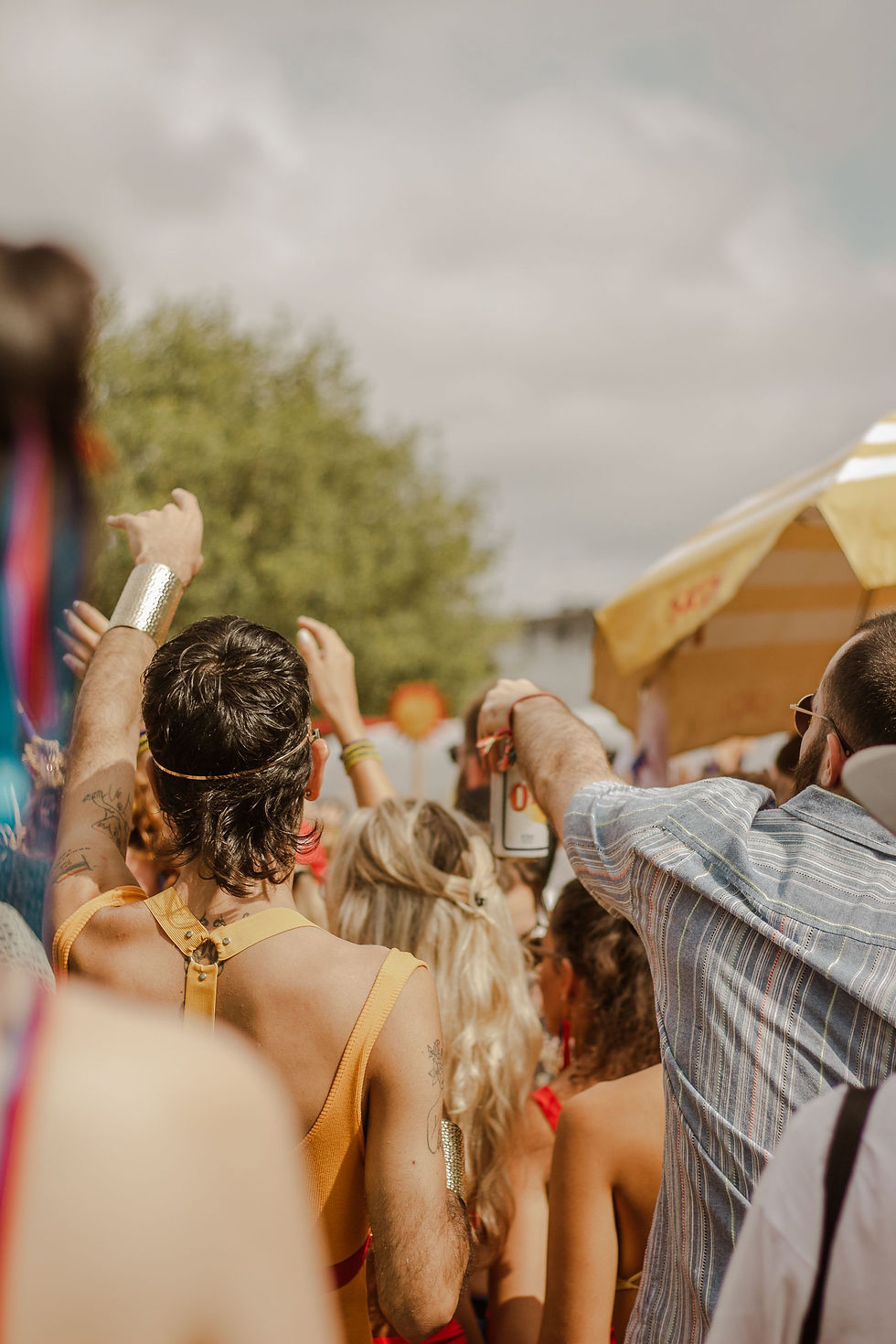 Crowd at an outdoor festival with raised arms, sunny sky, trees, and a yellow tent, creating a lively summer party mood