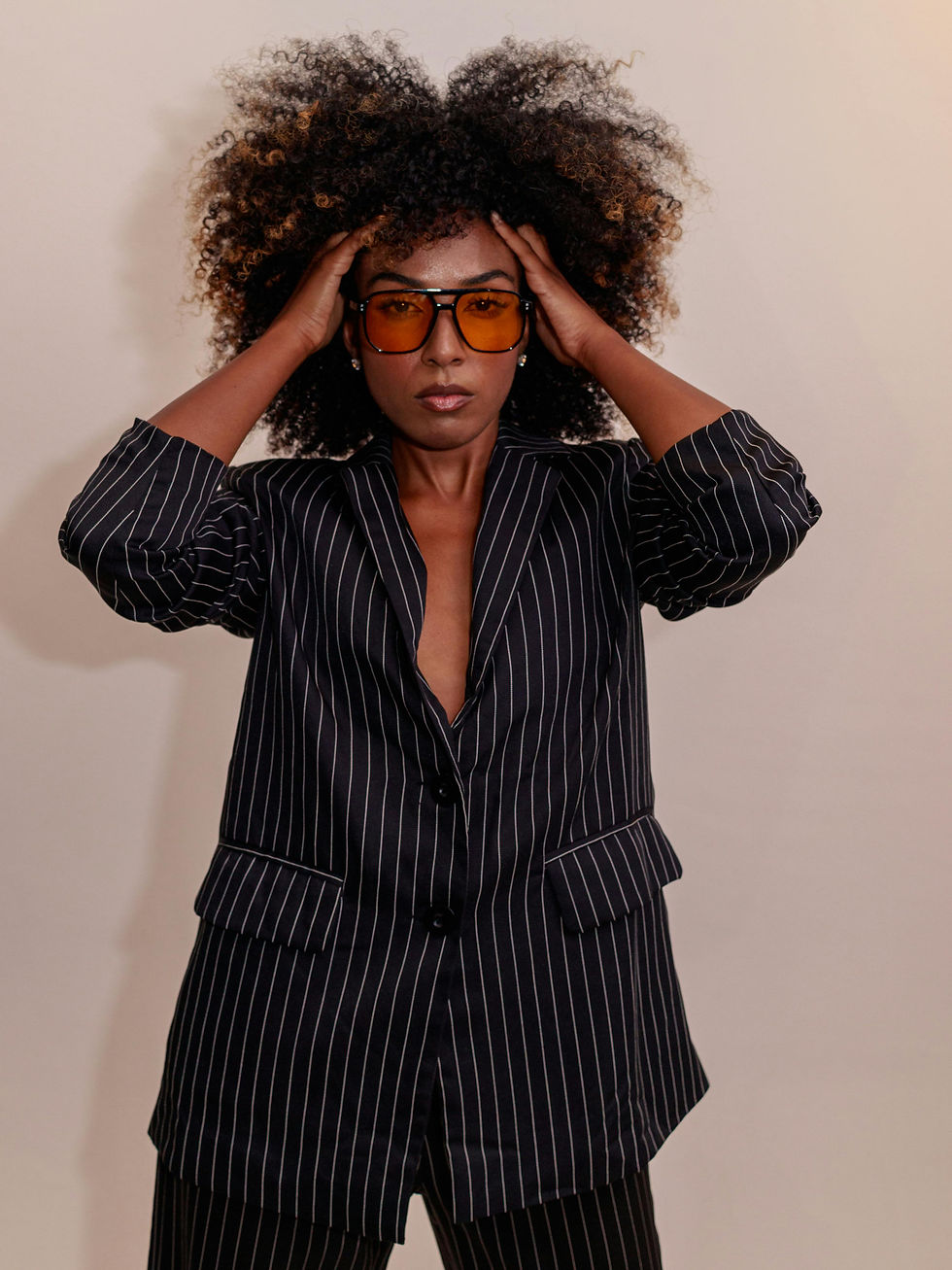 Woman in a black pinstripe vintage blazer and amber sunglasses poses against a plain wall, hands on her hair, with a confident look