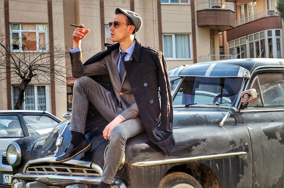Man wearing a vintage suit and vintage overcoat, sitting on the hood of a vintage car in a street