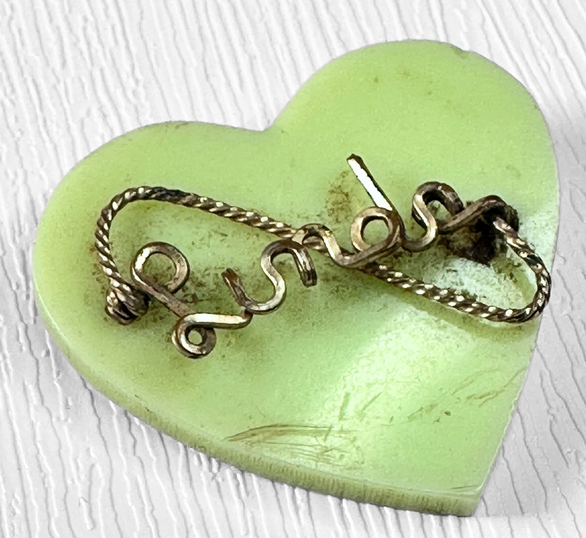 Green heart-shaped vintage brooch with bent metal wire clasp on a white textured surface, close-up.
