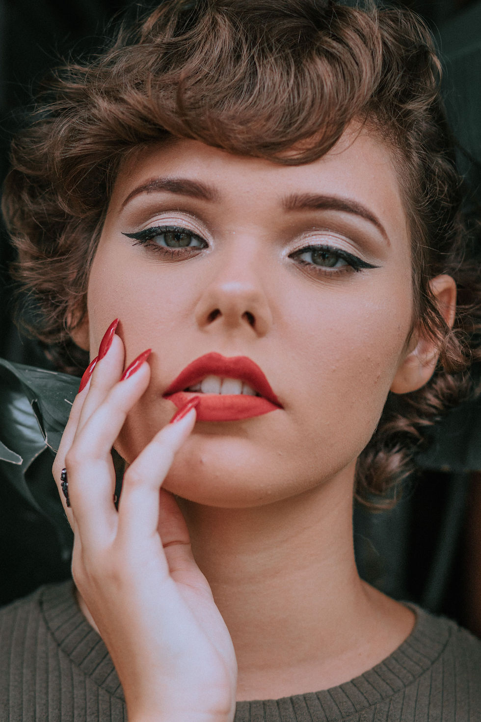 Close-up portrait of a woman with bold red lipstick and nails, dramatic eyeliner, and a hand on her cheek against a dark background