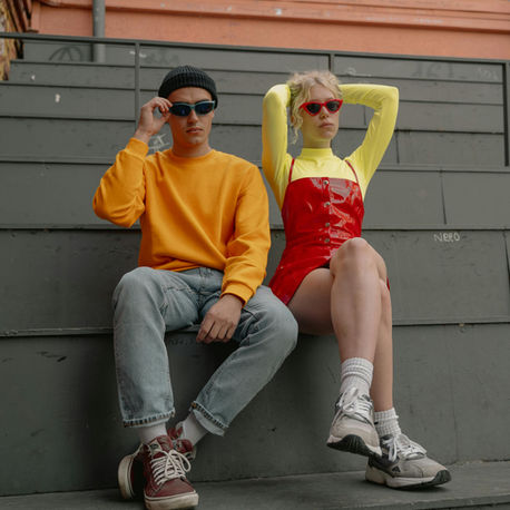 Two stylish young adults in 90s outfits and sunglasses sit on grey steps, posing bright orange, yellow, and red outfits against a brick wall.Decorative 1990s vintage clothing title card illustration