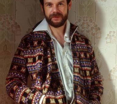 Bill Gibb stands indoors by patterned wallpaper, hands in pockets, wearing a colourful geometric jacket and white shirt.