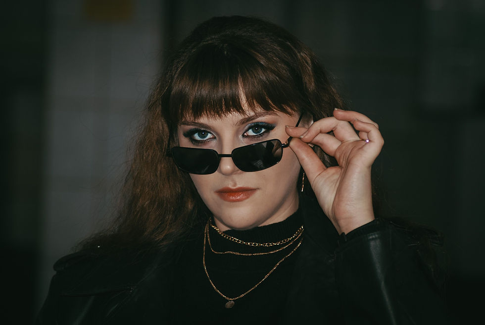Woman in black leather and layered gold necklaces lifts sunglasses, staring intensely against a dark background.