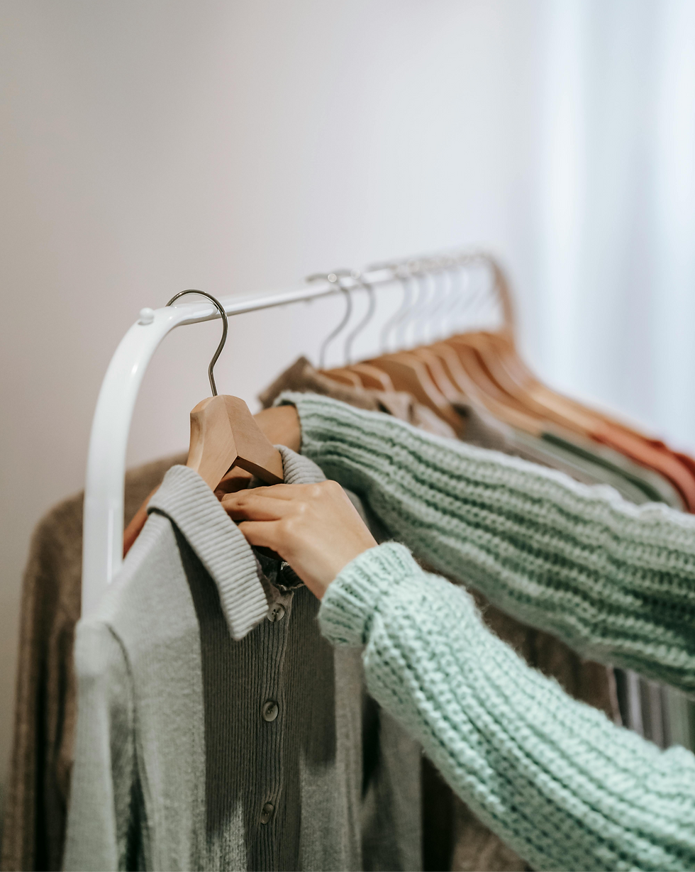 Hands browse knit sweaters on a clothing rack in a soft, minimalist shop, with muted gray and green fabrics in view.