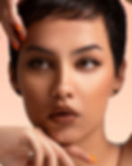 Close-up portrait of a woman with short dark hair, brown eyes, and orange nails framing her face against a peach background.