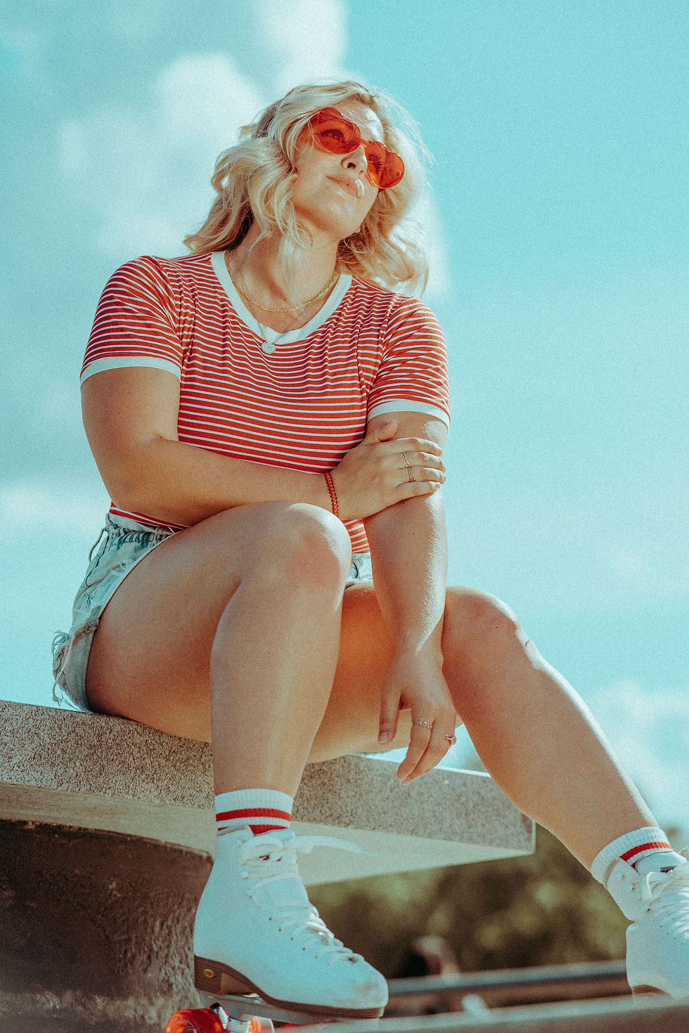 Blonde woman in red striped shirt and orange sunglasses sits on a ledge outdoors, wearing white retro rollerskates under a blue sky.