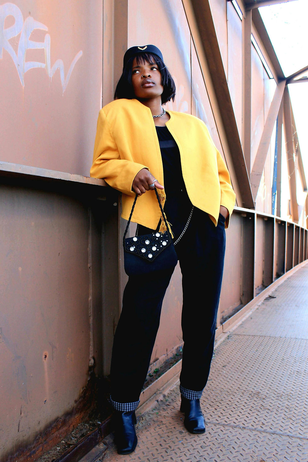 A person in a yellow vintage jacket and black outfit leans against a graffiti-marked metal wall, holding a decorative purse, on a bridge walkway.