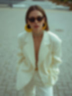 Woman in white suit with bold yellow earrings and sunglasses poses confidently on a paved street. Urban background, chic fashion vibe.