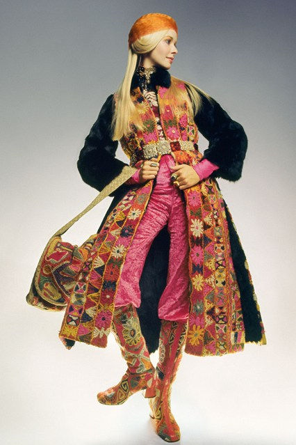 Fashion model posing in a Thea Porter ensemble - colourful bohemian coat, pink pants and boots, with a patterned bag, against a grey studio background.