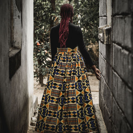 Woman with red braids stands in a narrow alley between concrete walls, wearing a black top and colourful patterned maxi skirt.