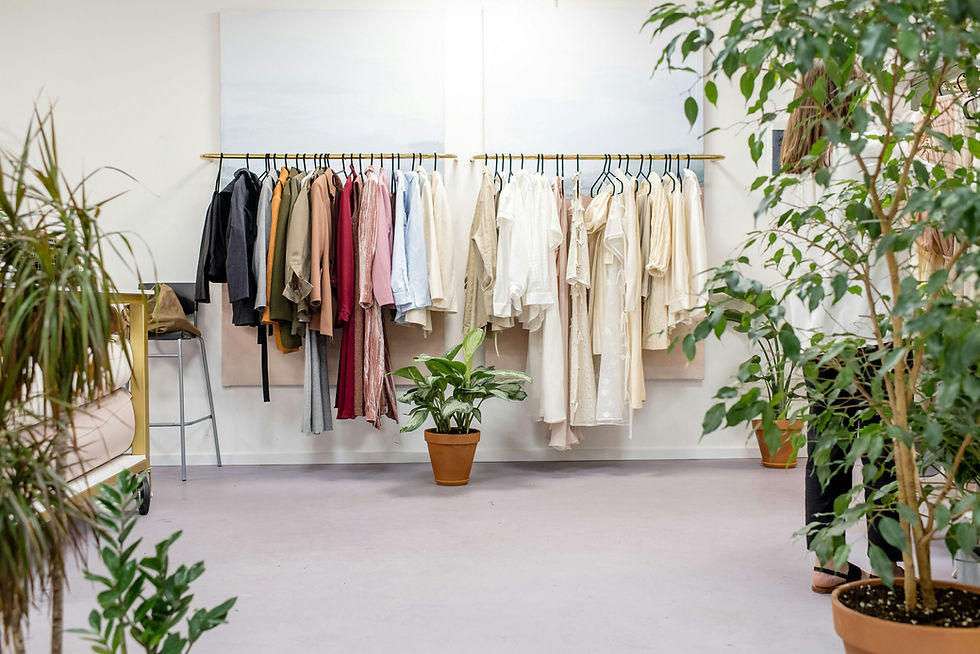Minimalist boutique with neutral and colourful clothes on racks, potted plants, and two people browsing in the background.