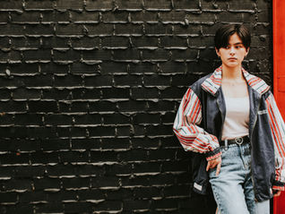 Young person in striped vintage tracksuit jacket leans against a black brick wall beside a red door, looking pensive.