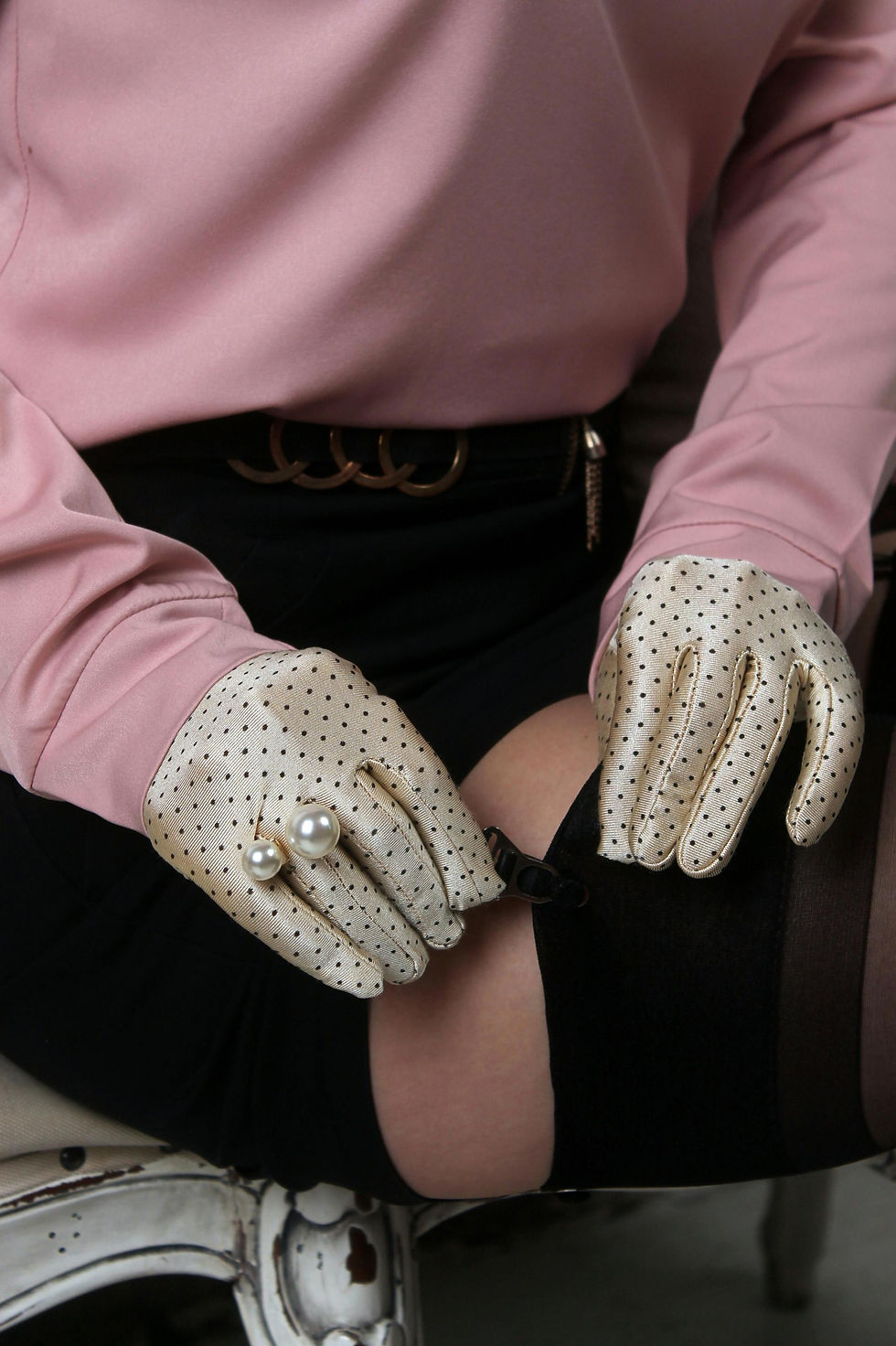 A person wearing a pink blouse and polka dot gloves clips a black stocking in place. A pearl ring adorns their hand. Elegant mood.