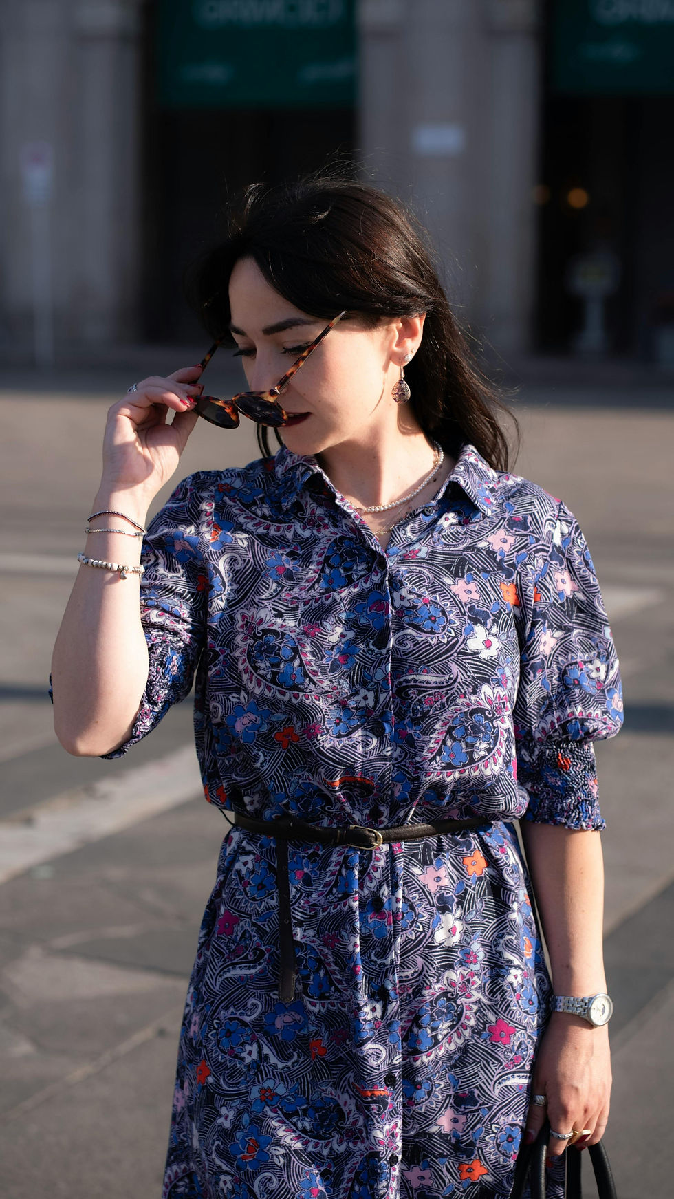 Woman in a floral vintage dress adjusts sunglasses, standing on a street. Background is blurred. She's wearing jewelry and appears calm.
