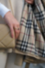 Close-up of a hand with gold rings holding a quilted handbag, beside a Burberry plaid scarf and sweater in a soft fashion shot