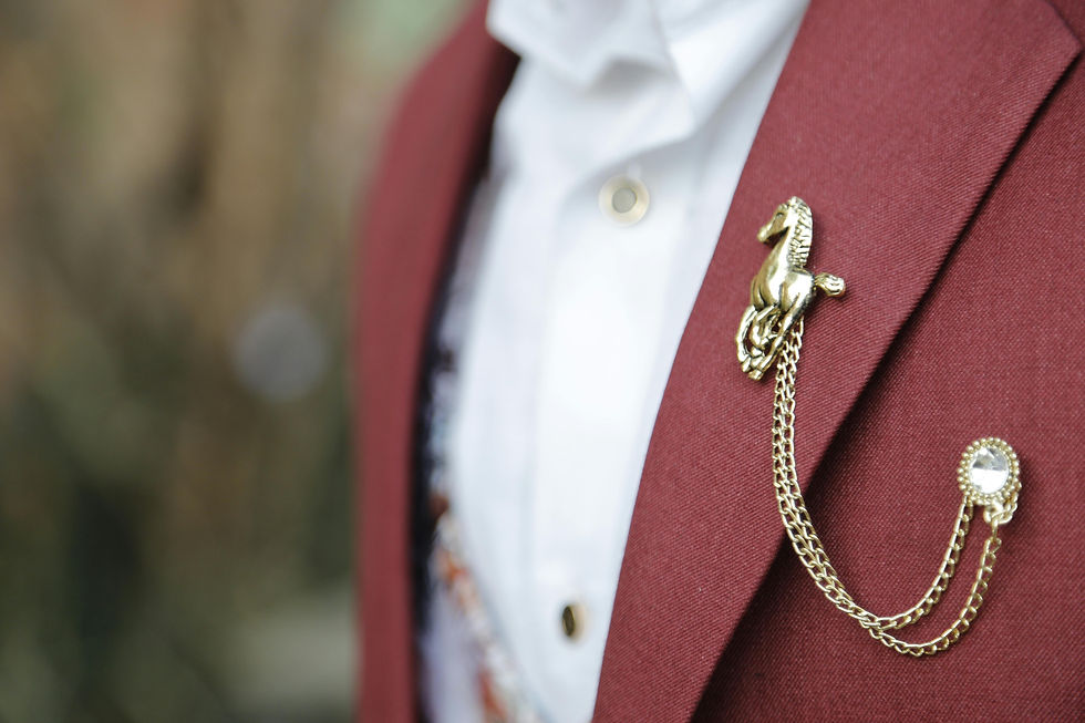 Close-up of a maroon suit with a gold horse vintage brooch and chain. The white shirt underneath creates a formal look. Blurred background.