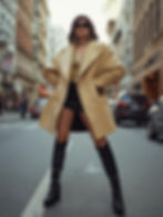 Woman in a beige coat, leopard print top, black shorts, and boots stands confidently on a city street, wearing sunglasses and earrings.