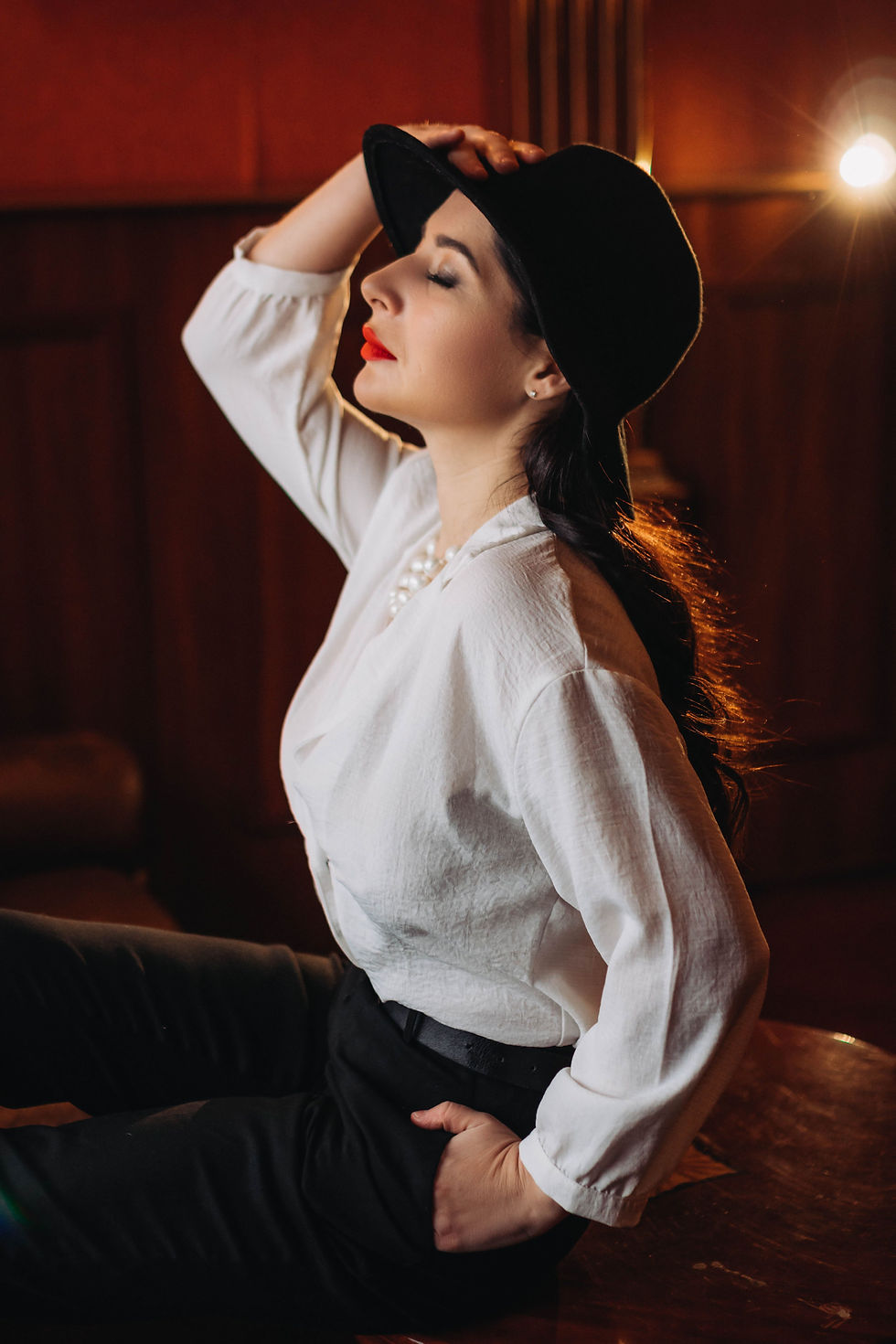 Woman in black hat and white blouse poses in warm studio light, eyes closed, red lips, seated against a dark background.