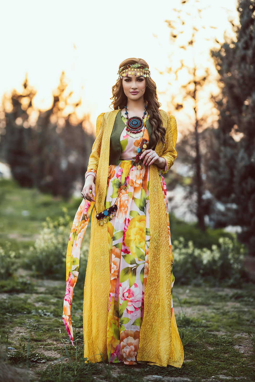Woman in a colorful floral dress and yellow shawl stands outdoors at sunset, wearing a beaded headband and necklace, surrounded by trees.