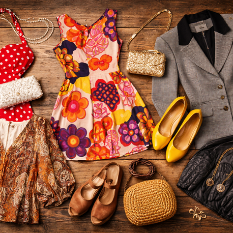 Vintage clothing and accessories on a wooden floor, featuring colorful dresses, a gray blazer, yellow heels, woven bag, and pearl necklace.