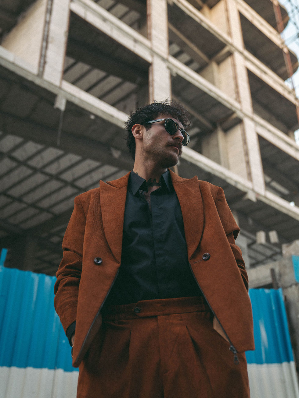Man in a rust-coloured vintage suit and sunglasses stands under a concrete construction frame, with blue fencing behind him, looking confident.
